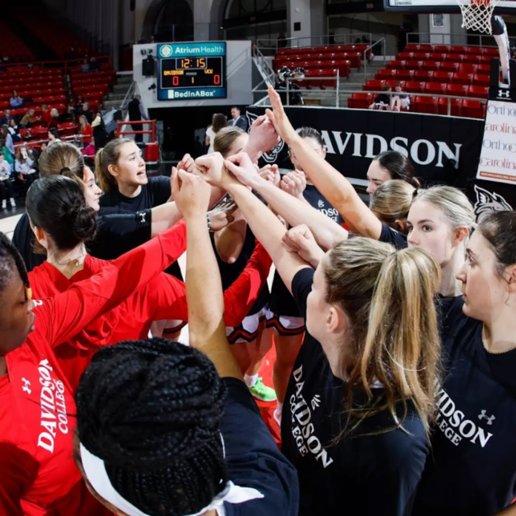 Davidson Women's basketball team in a huddle with their hands together in the middle on their home court.