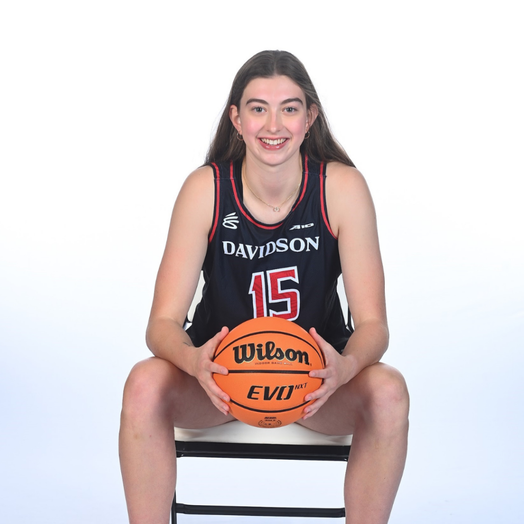 Eliza Buerk holding a basketball and leaning forward on a chair. She's wearing her Davidson basketball uniform. 