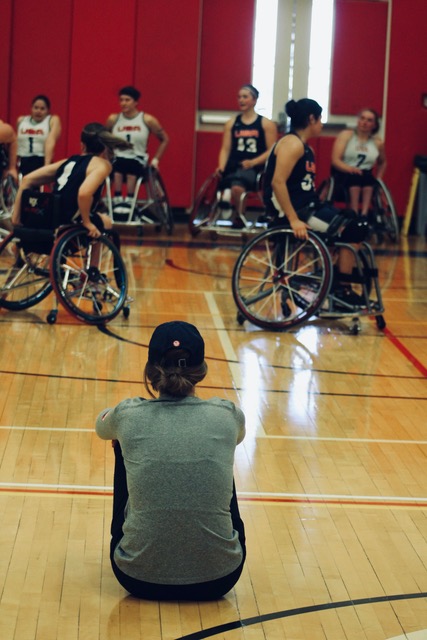 Abby Dunkin sitting on the ground of an indoor gym watching Team USA players compete in Wheelchair Basketball