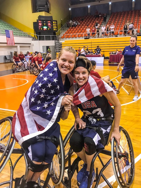 Abby and another Wheelchair Basketball athlete posing together with an American flag around their shoulders. They are inside an indoor gym.