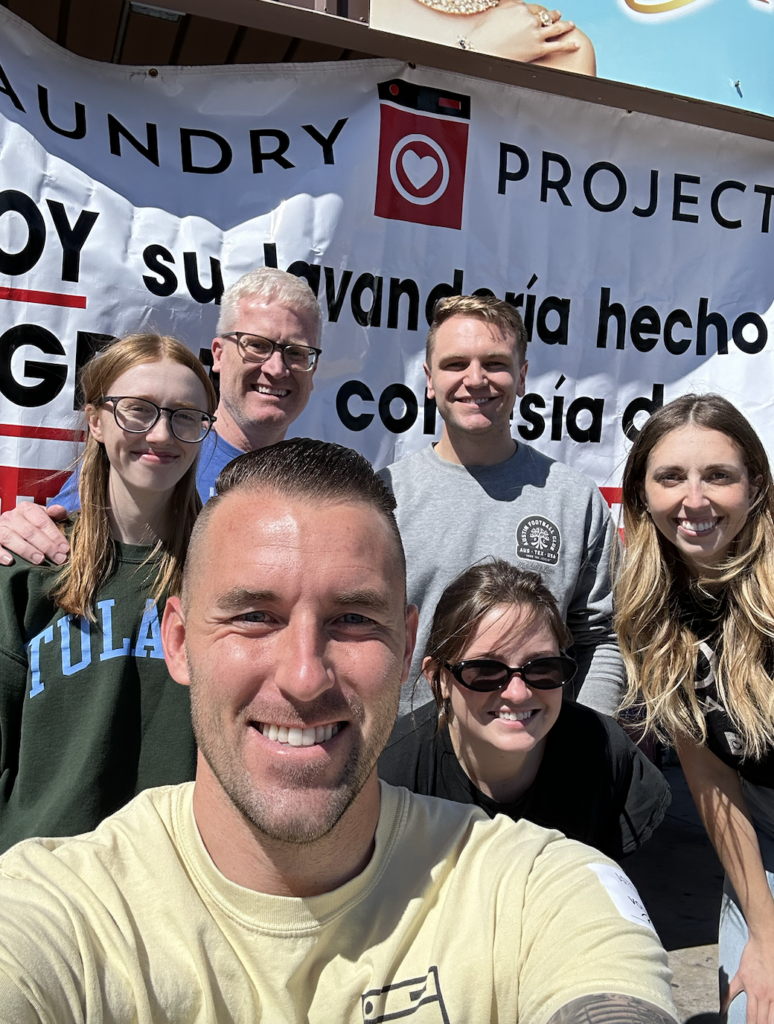 Brad takes a selfie with others, including Ashley, in the background in front of a sign for the Laundry Project. 