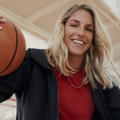 Headshot of Elena Delle Donne. She is smiling, wearing a black blazer, a red shirt and holding a basketball.