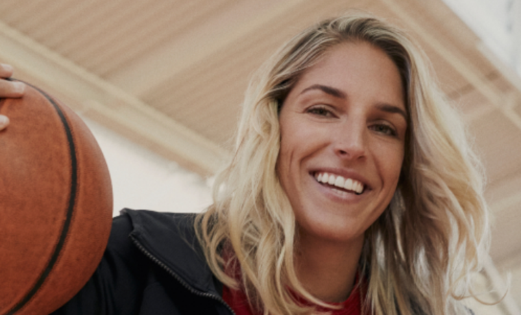 Headshot of Elena Delle Donne. She is smiling, wearing a black blazer, a red shirt and holding a basketball.